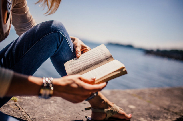 A picture of a lady reading a book in the sunshine by the water