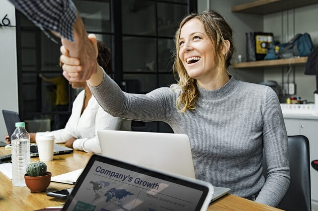 Picture of a lady at her desk with a laptop smiling happily as someone approaches to shake her hand. Show two people engaging in a happy friendly relationship.