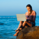 Woman sitting on a rock with a laptop close to the sea