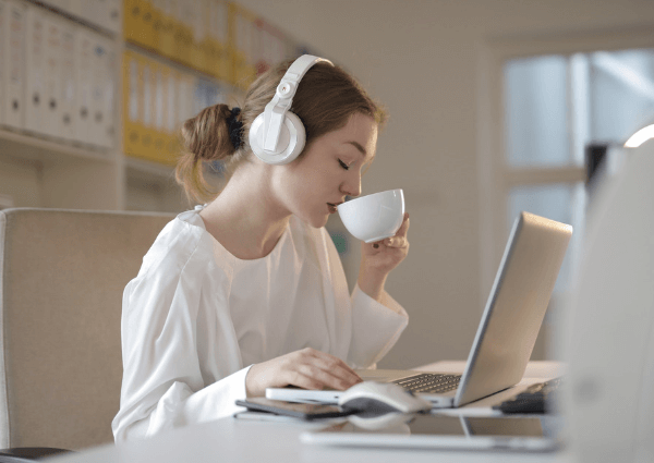 Image of a woman wearing headphones and drinking tea while working on her laptop