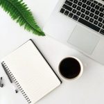 Picture of a workspace desk with a green plant, a laptop, notepad and cup of coffee.