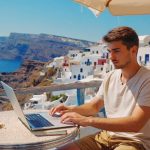 A digital nomad sitting at a table working on his laptop in the sun with a holiday setting in the background